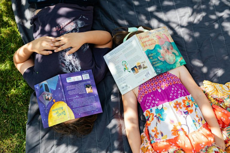 Two children lying on a blanket outdoors with books covering their faces, enjoying a sunny day.